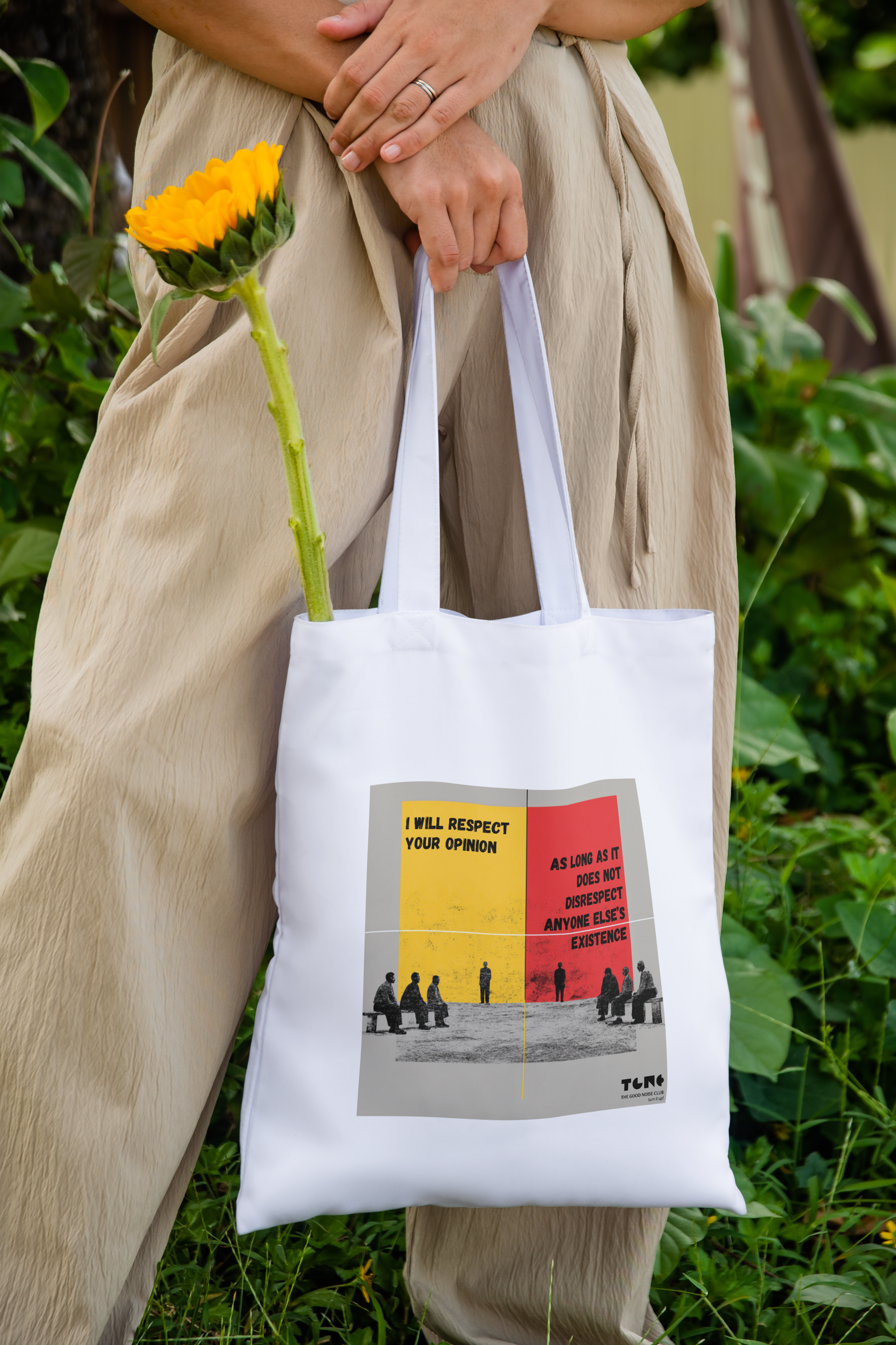Person holding a white tote bag with a graphic design, standing outdoors with greenery.