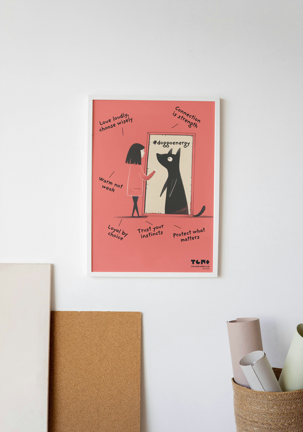 Framed artwork with a cat and person illustration on a pink background, leaning against a white wall.