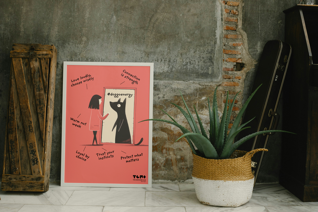 Poster with a black cat illustration on a pink background, placed against a textured wall with a plant and wooden crate.