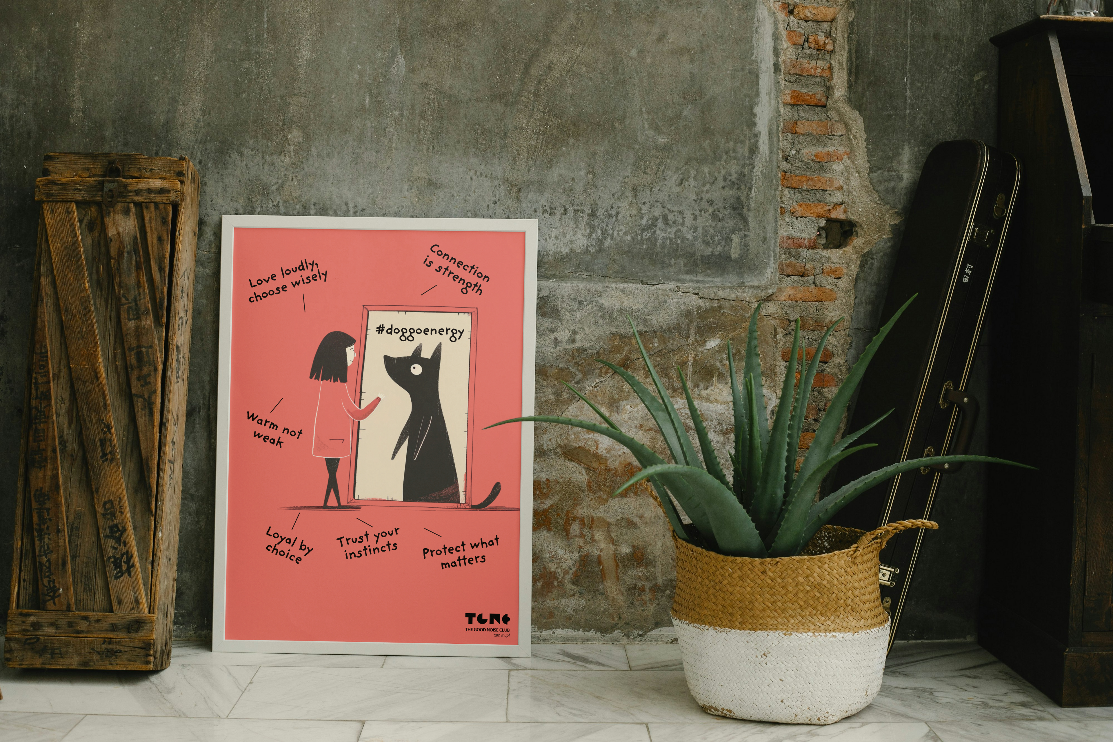 Poster with a black cat illustration on a pink background, placed against a textured wall with a plant and wooden crate.