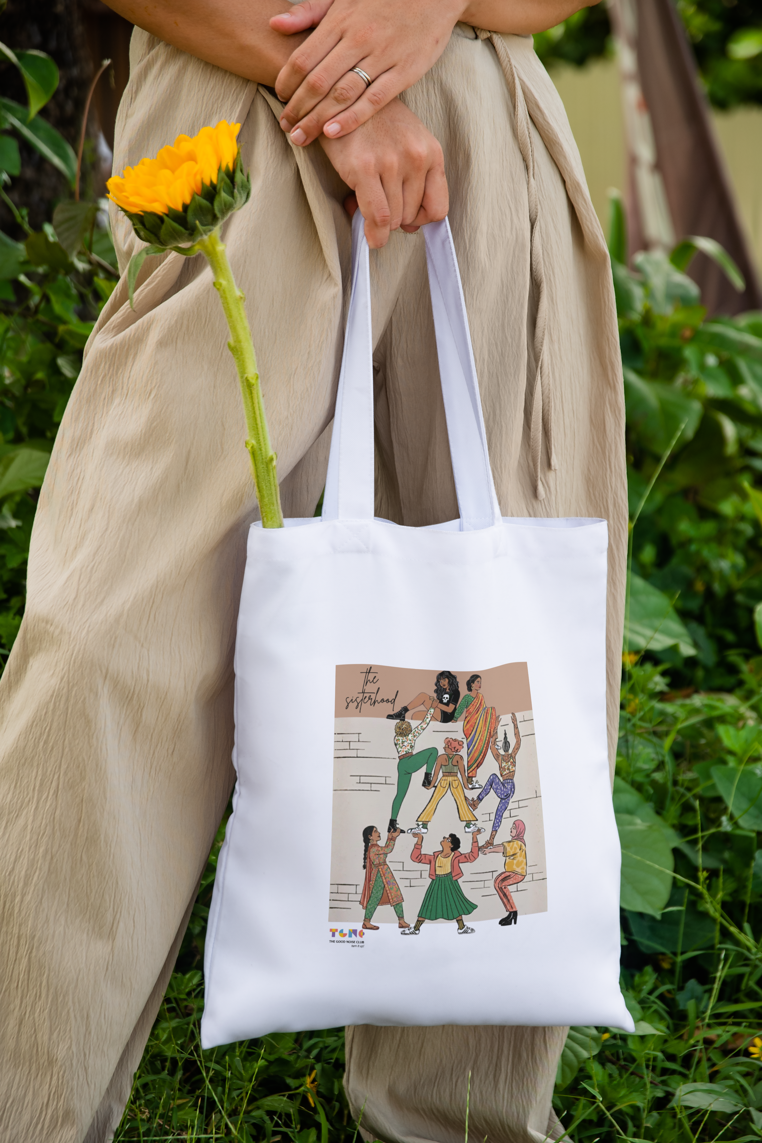 Person holding a white tote bag with a colorful design, standing outdoors with greenery.