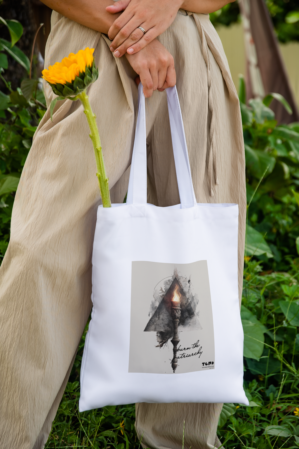 Person holding a white tote bag with a design and a sunflower in a natural setting