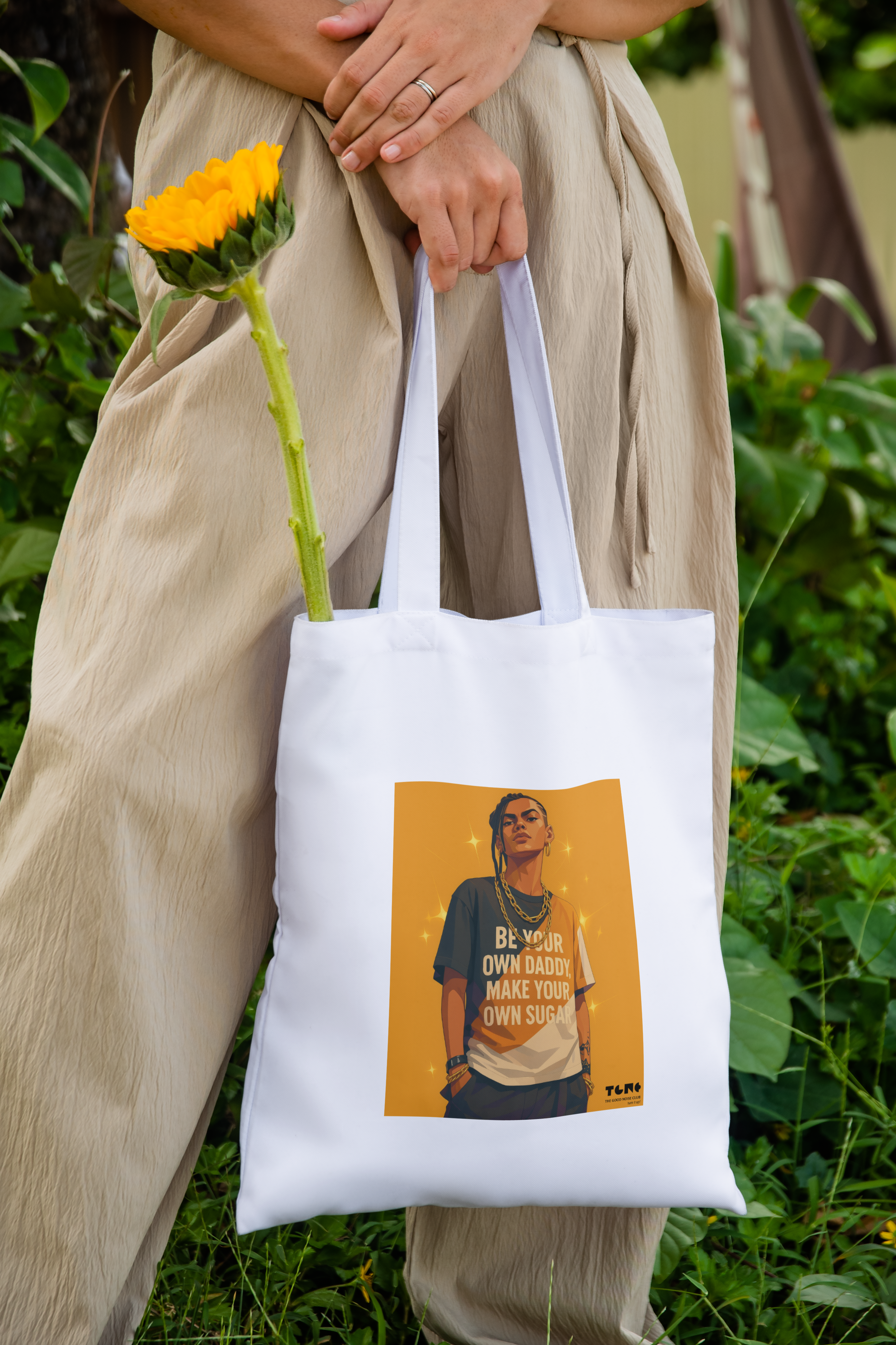Person holding a white tote bag with a printed design, standing outdoors with greenery in the background.