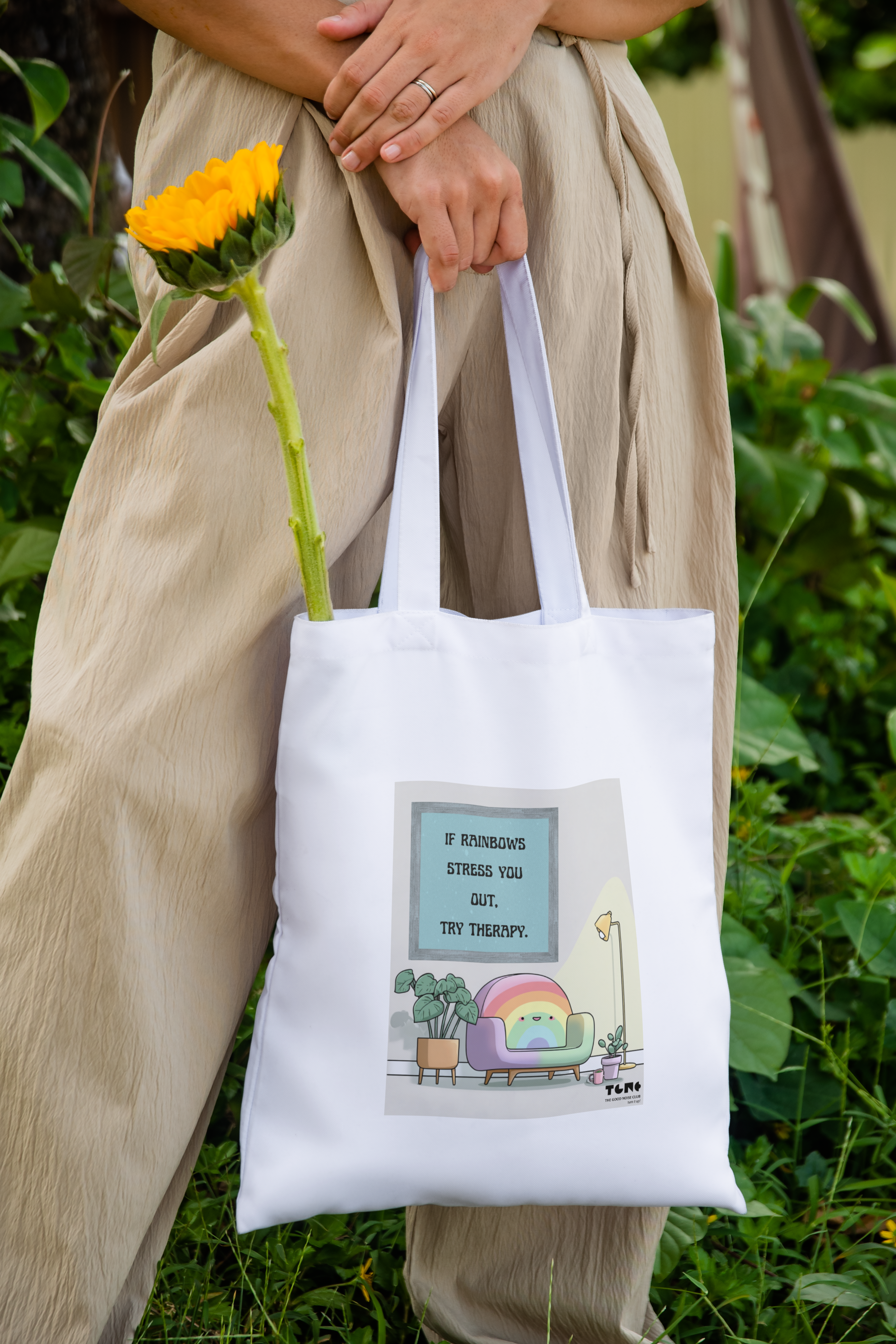 Person holding a white tote bag with a colorful design, standing outdoors with greenery.