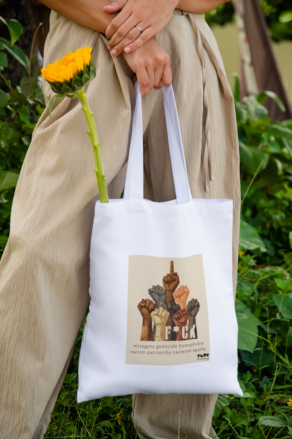 Person holding a white tote bag with a graphic design, standing outdoors with greenery.