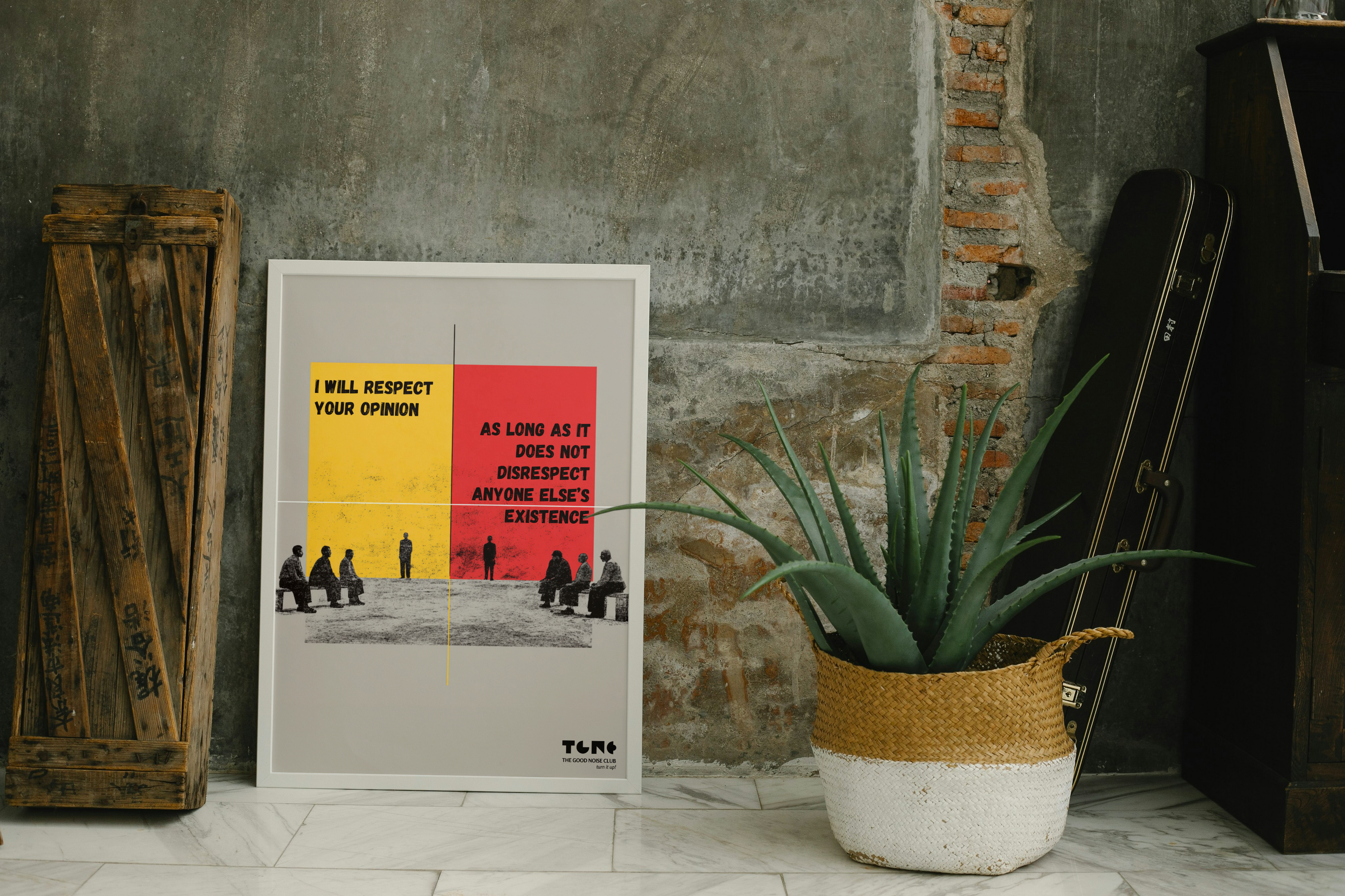 Framed poster with text on a wall next to a plant and wooden crate.