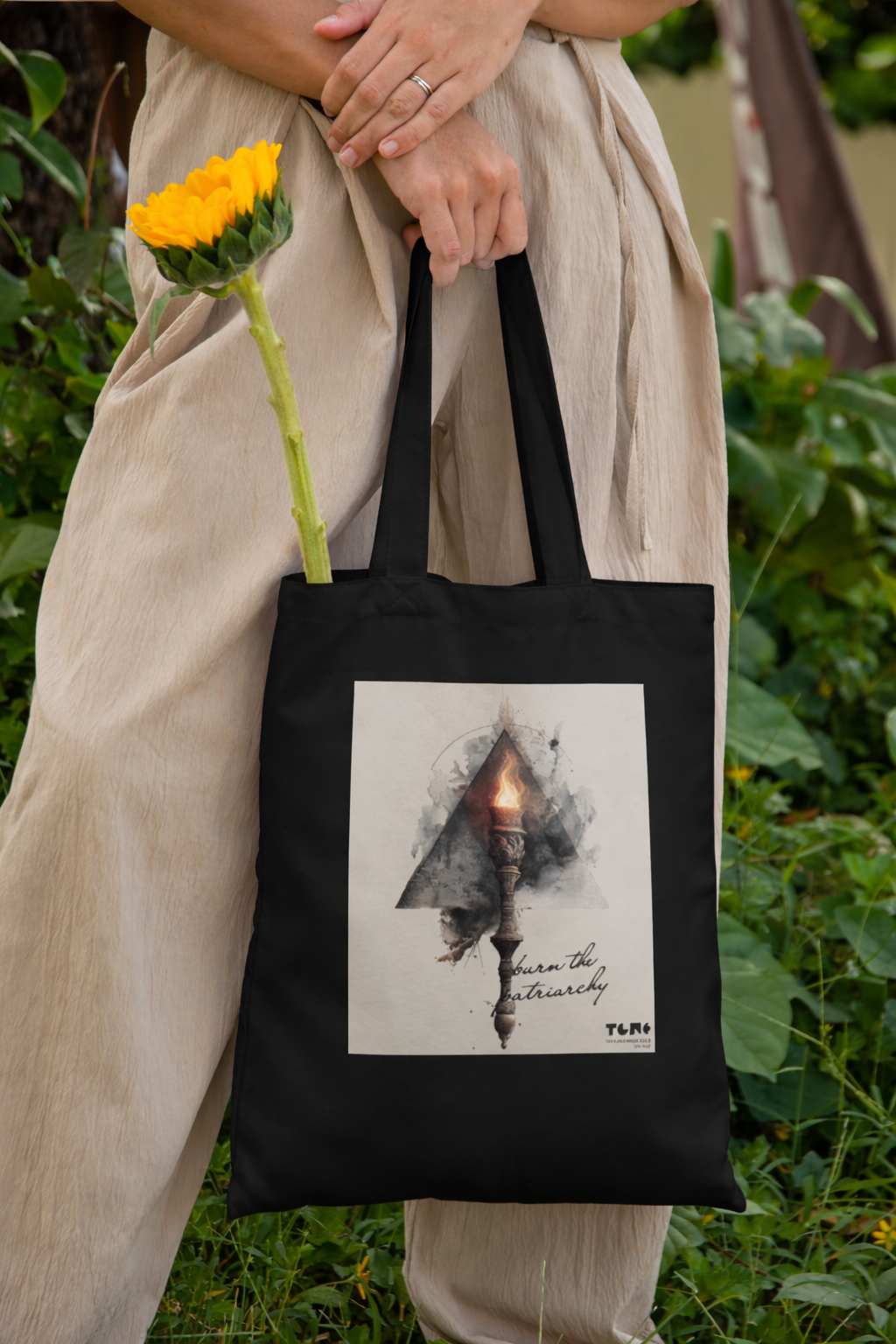 Person holding a black tote bag with a design and a sunflower in a natural setting