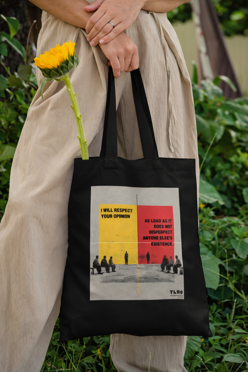 Person holding a black tote bag with a graphic design and text, standing outdoors.