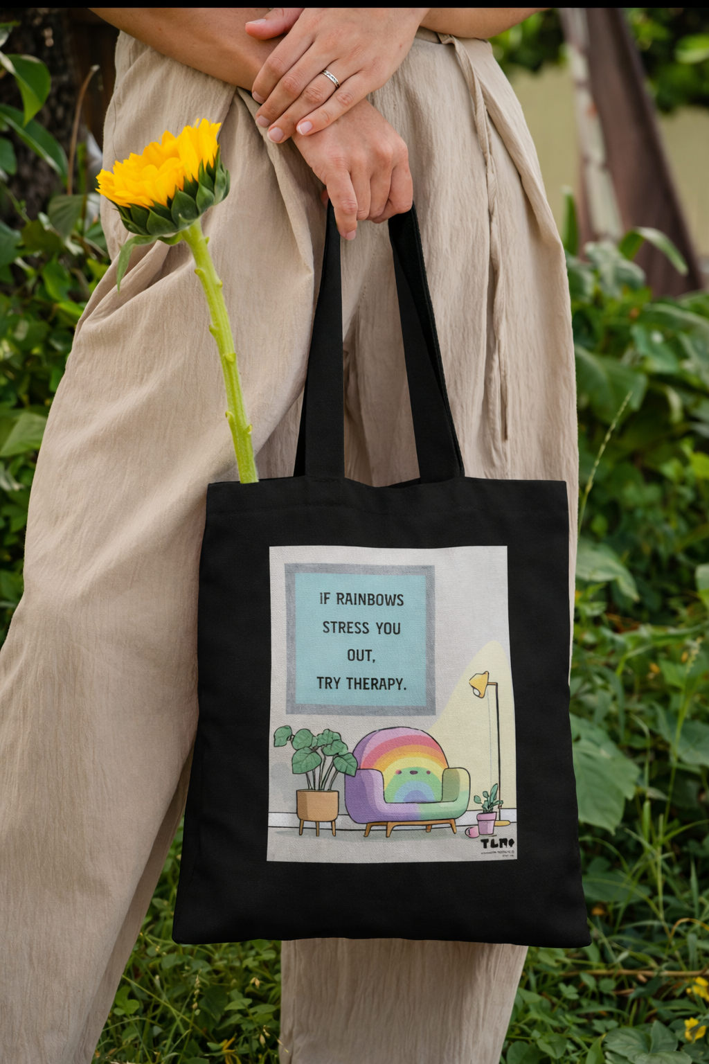 Person holding a black tote bag with a colorful design and text, standing outdoors.