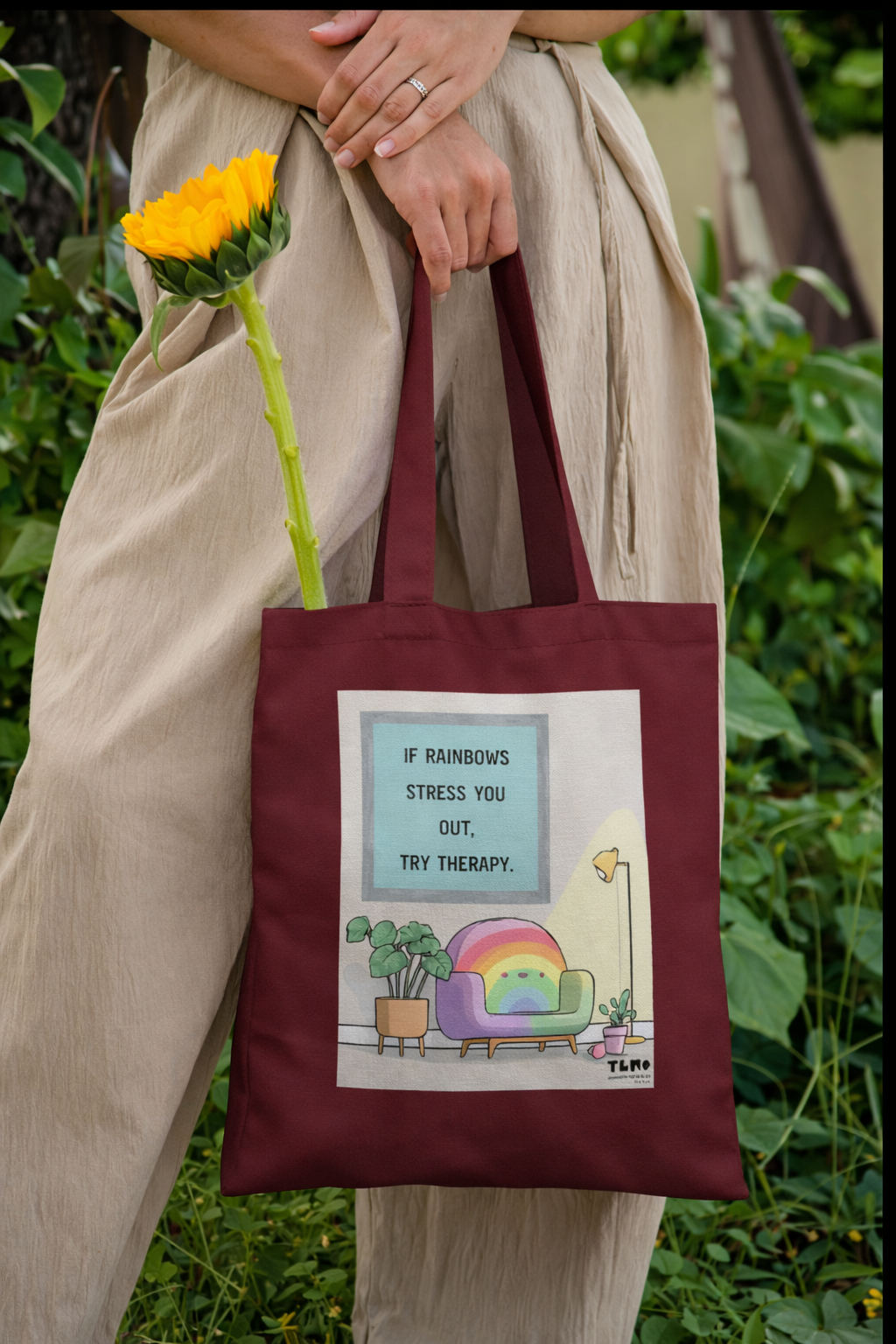 Person holding a maroon tote bag with a colorful design and text, outdoors.