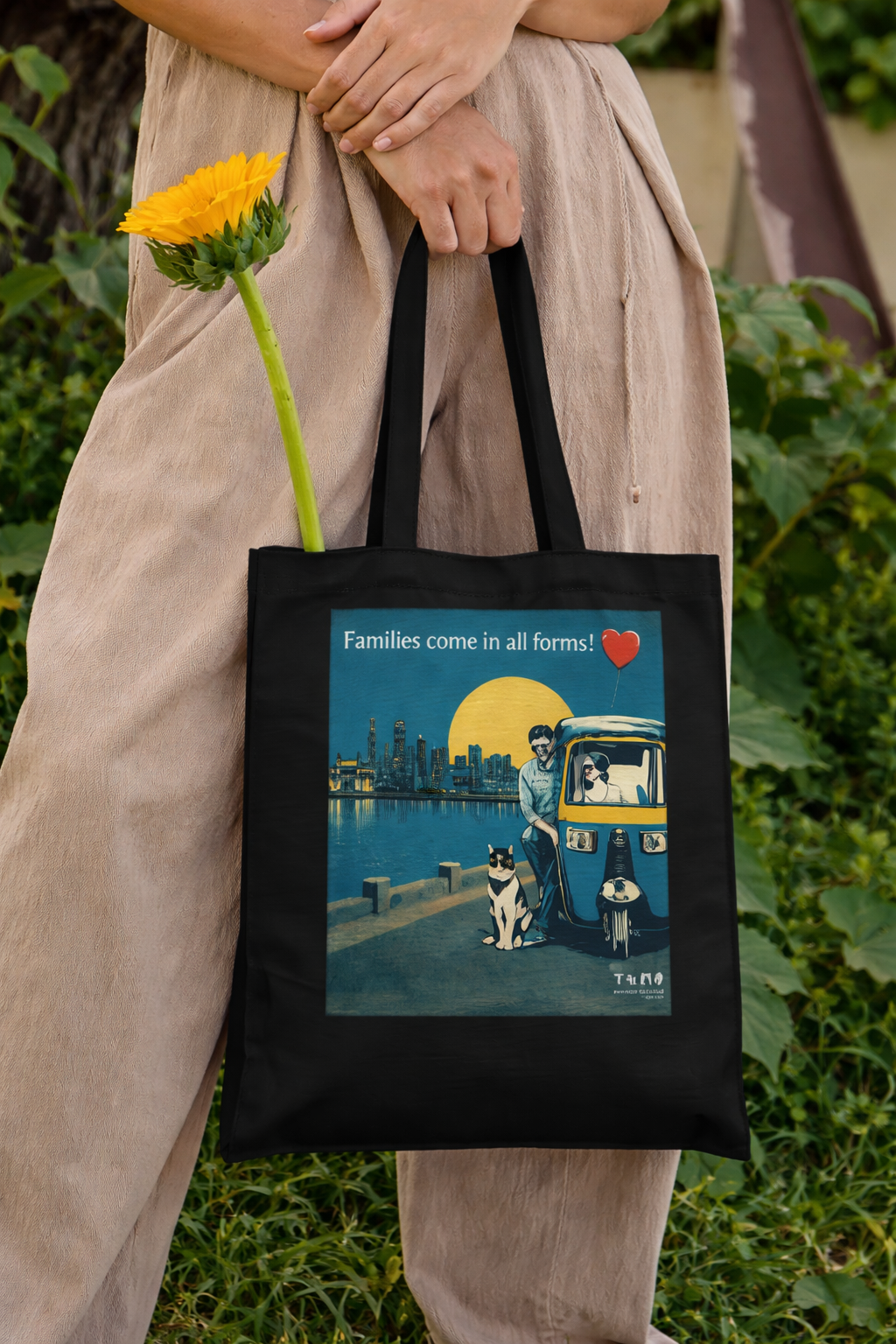 Person holding a black tote bag with a colorful design featuring a couple and two dogs, set against a green outdoor background.