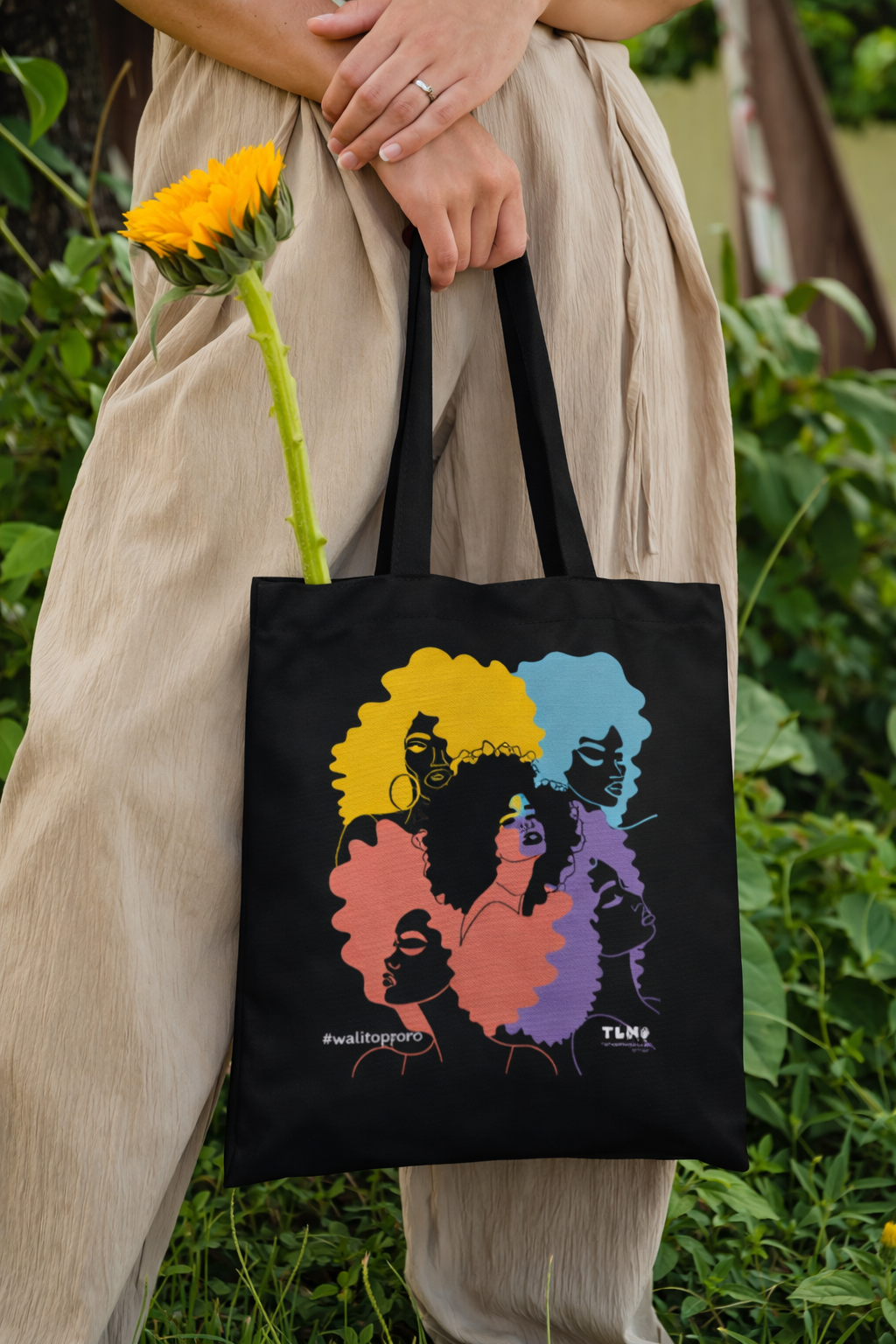 Person holding a black tote bag with colorful design and a sunflower, standing in a natural setting.