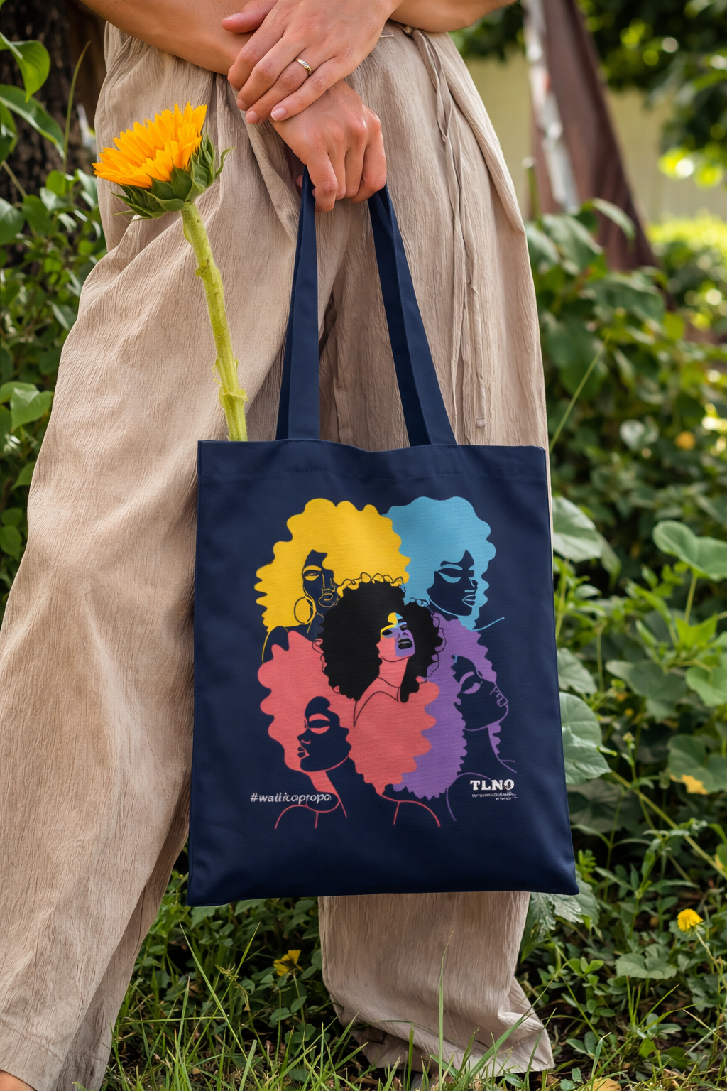 Person holding a tote bag with colorful designs of women's faces in a garden setting