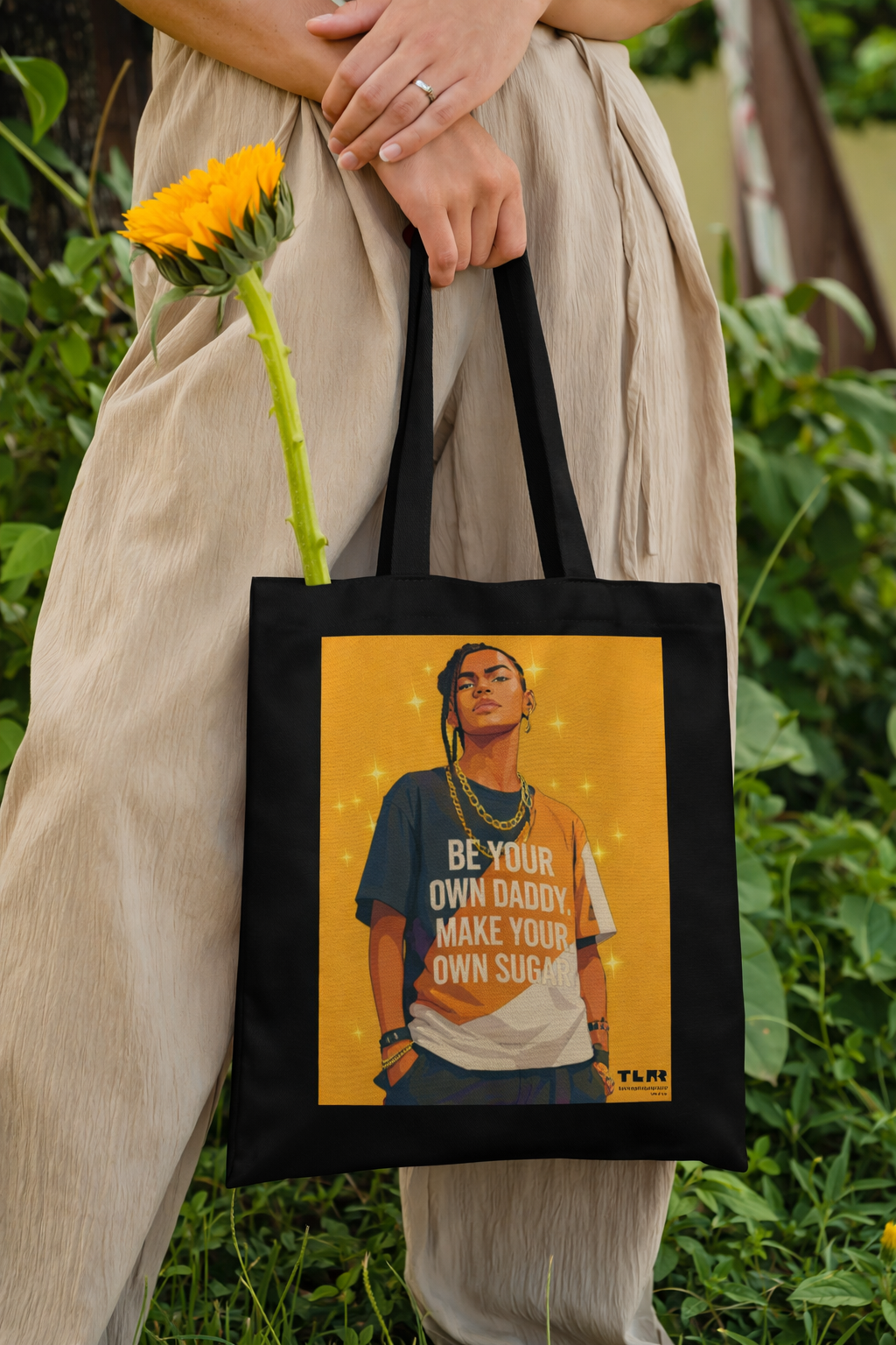 Person holding a tote bag with a graphic design and text, standing outdoors.