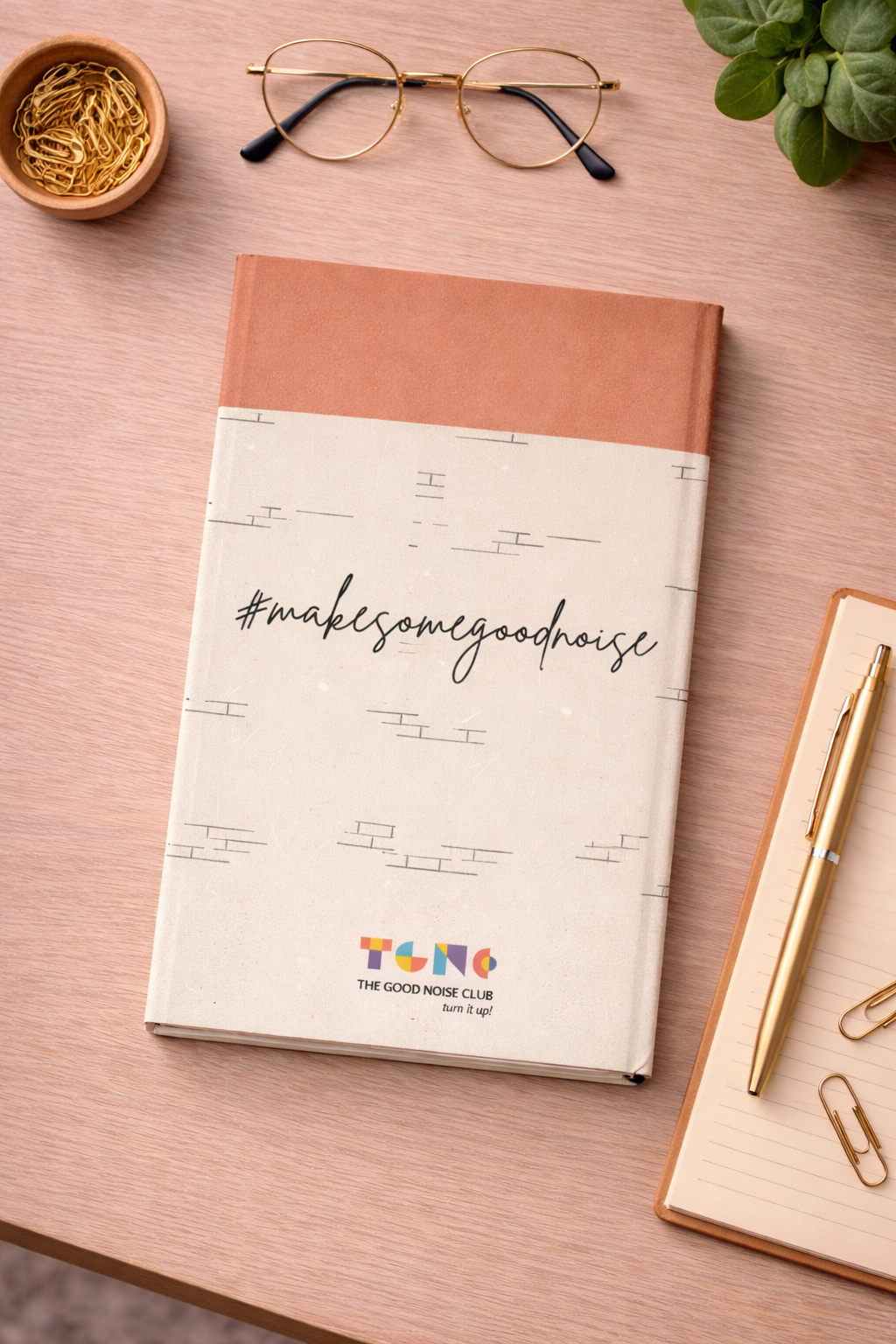 Notebook with 'make some good noise' text on a desk with glasses, pen, and plant
