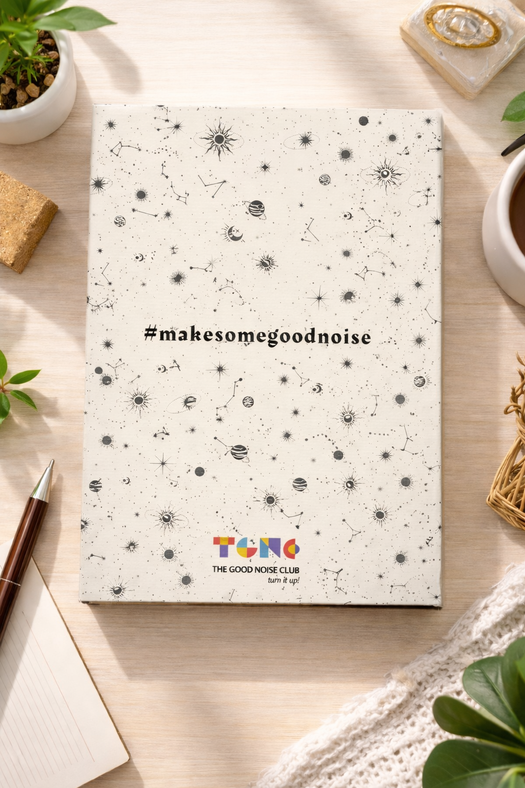 Notebook with '#makesomegoodnoise' text on a desk with plants and a pen.