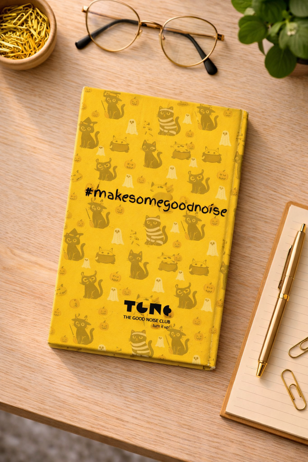Yellow notebook with cat pattern and 'makesomegoodnoise' text on a wooden surface with glasses and a pen.