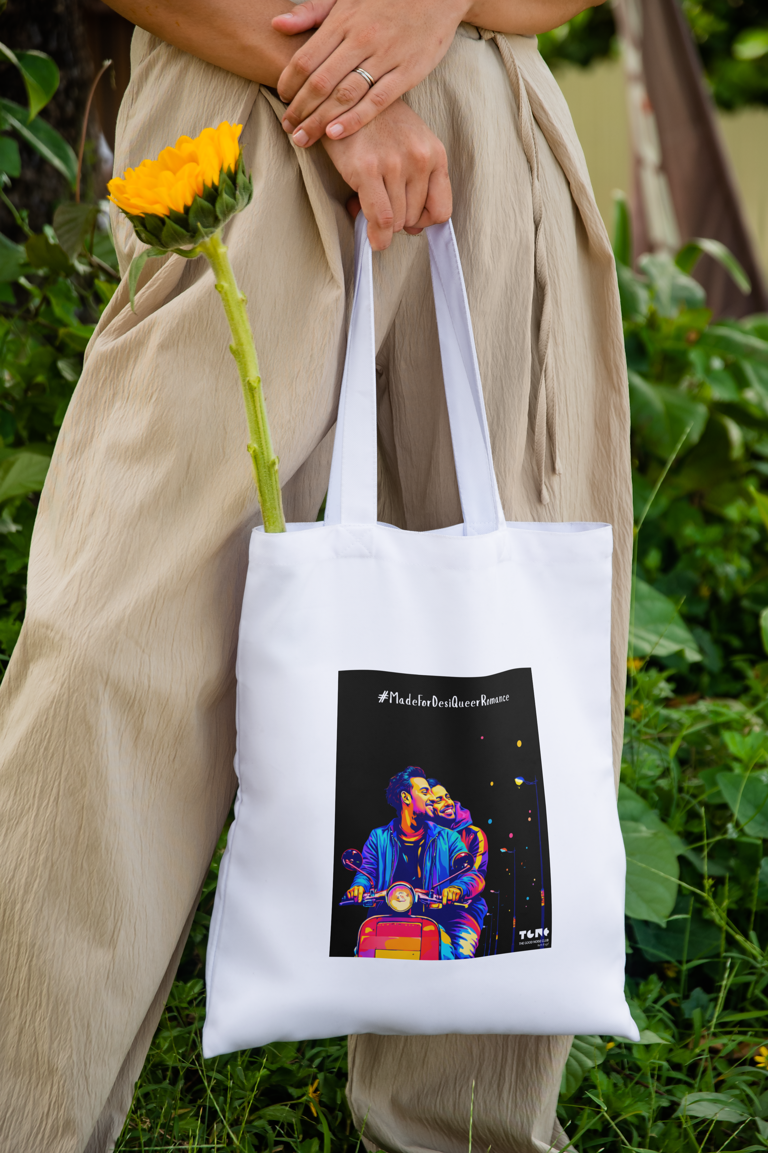 Person holding a white tote bag with a colorful graphic, standing outdoors with greenery.