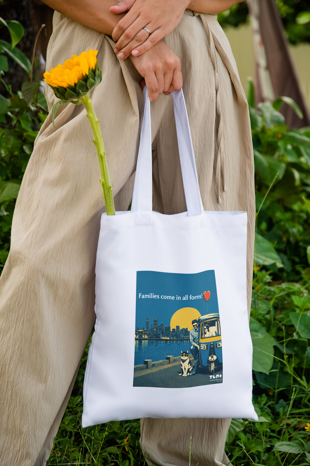 Person holding a white tote bag with a design of a cityscape and two dogs, standing outdoors with greenery.