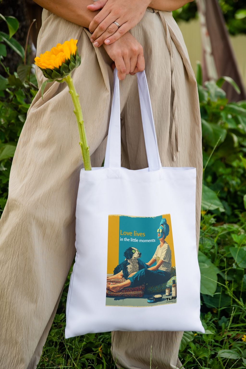 Person holding a white tote bag with a design, standing outdoors with greenery.