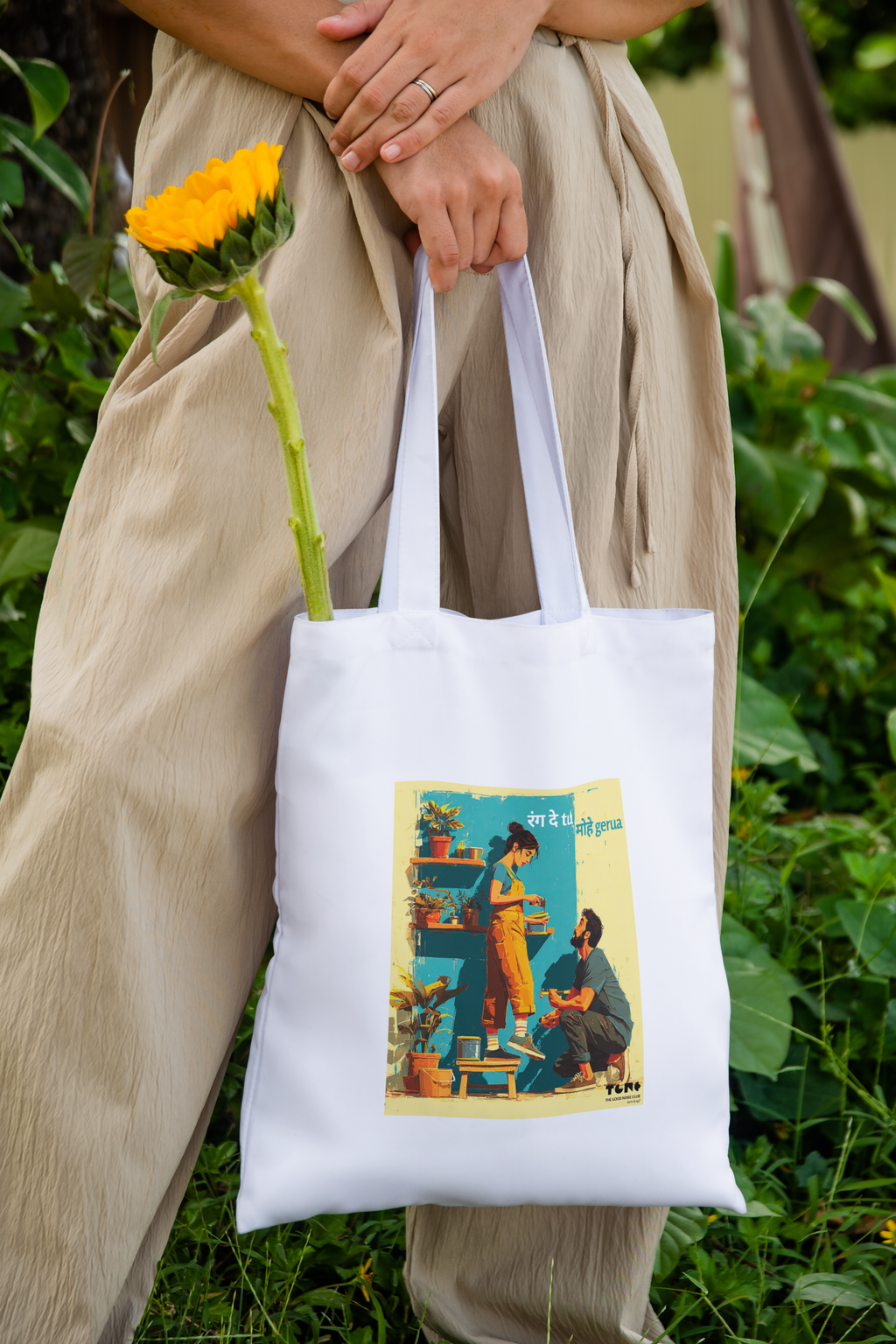 Person holding a white tote bag with a design, standing outdoors with greenery.