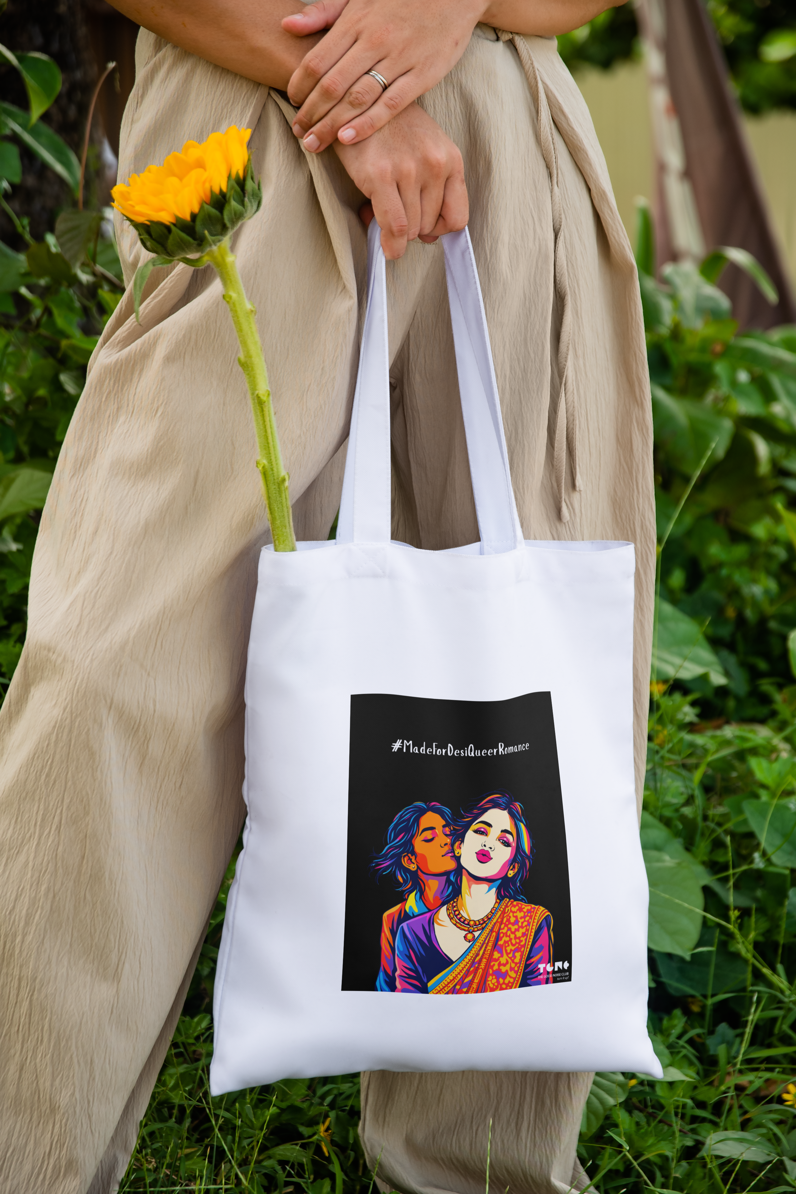 Person holding a white tote bag with a colorful design of two people, one holding a yellow flower, against a green outdoor background.