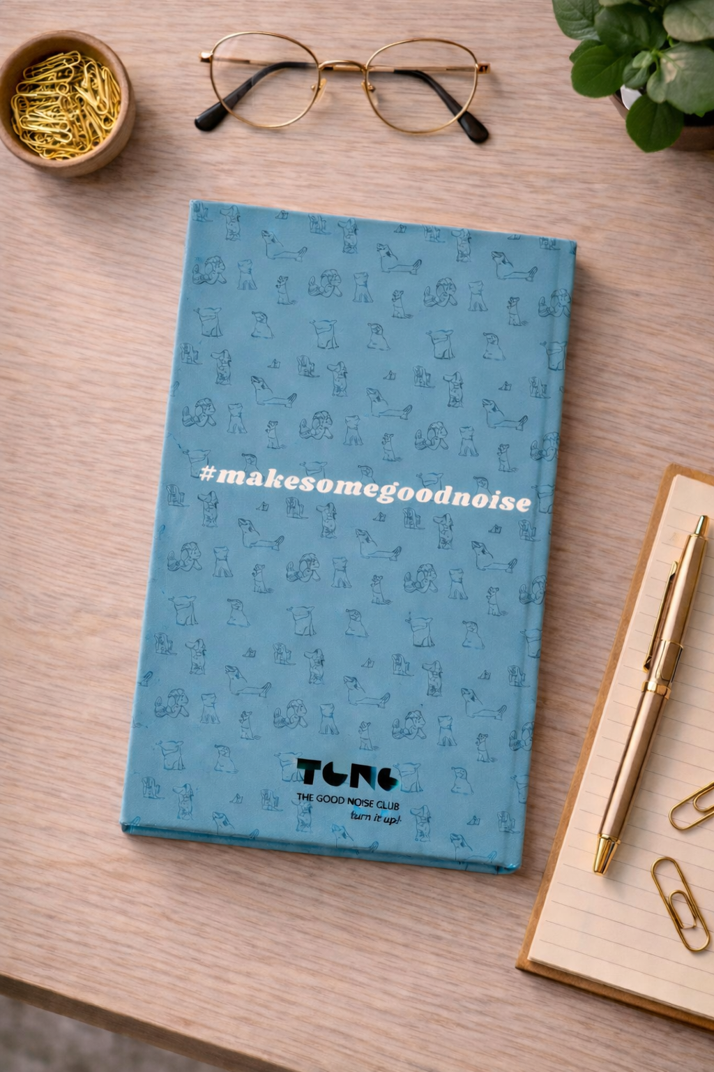 Blue notebook with 'makesomegoodnoise' text on a wooden surface with glasses, pen, and paper clips.