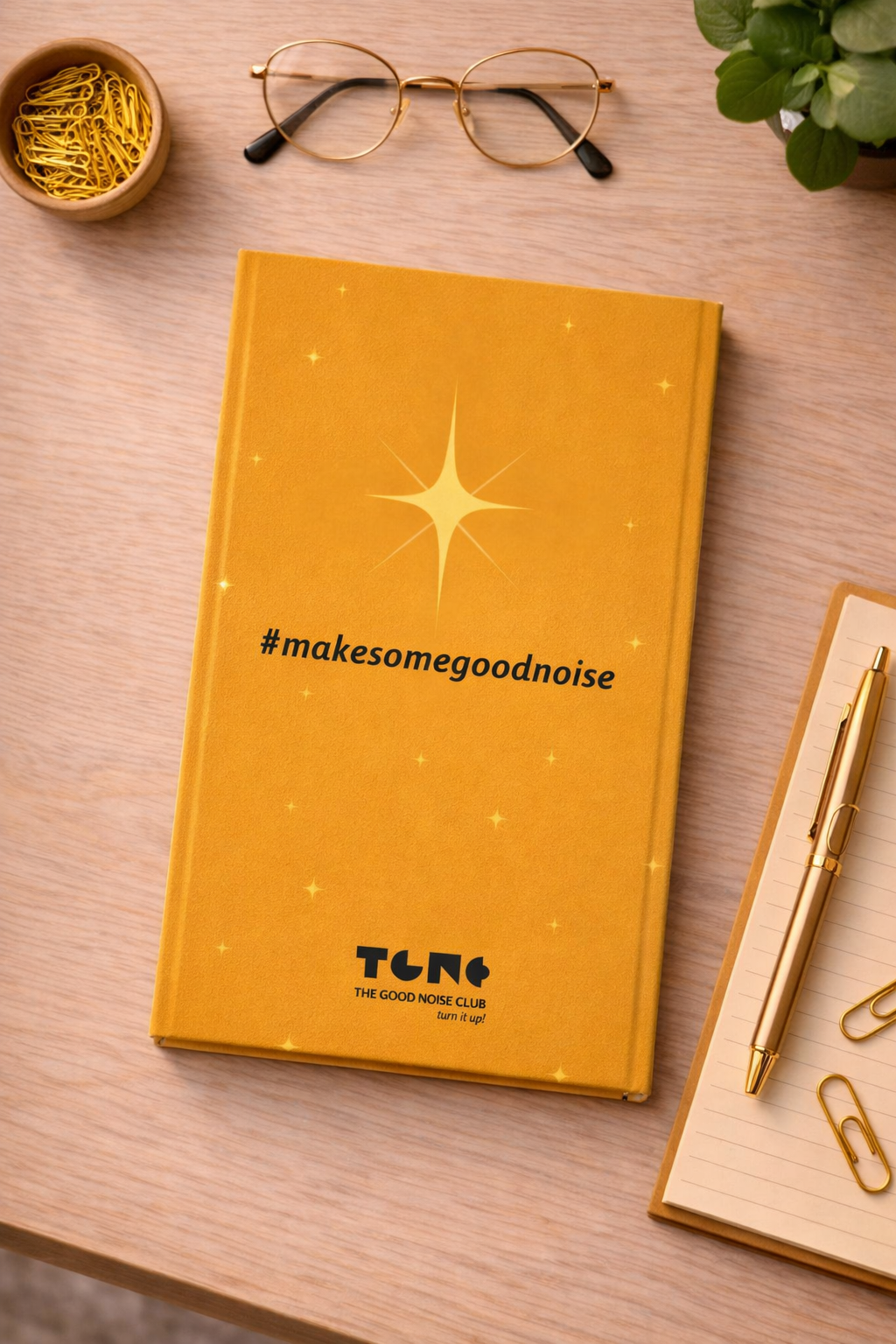 Yellow notebook with '#makesomegoodnoise' text on a wooden surface with glasses and a pen.